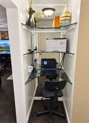 Un espace de travail compact aménagé dans un coin de la maison, comprenant un bureau en verre, un ordinateur portable, une imprimante et une chaise.