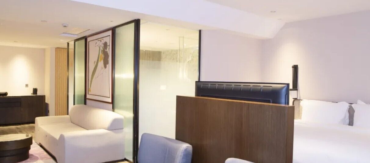 A modern hotel room with a bed, a seating area with a chair, a desk, and a large window.