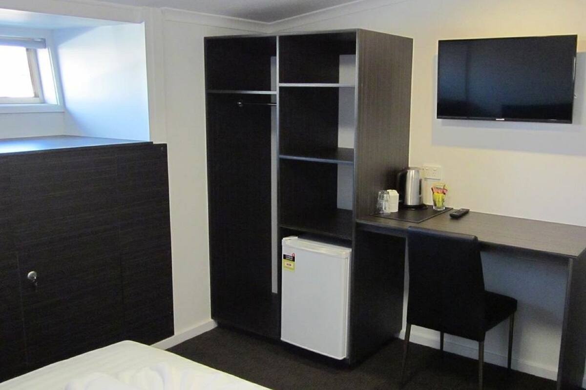 A compact room with a bed, a desk, a chair, a small refrigerator, a TV, and a window.
