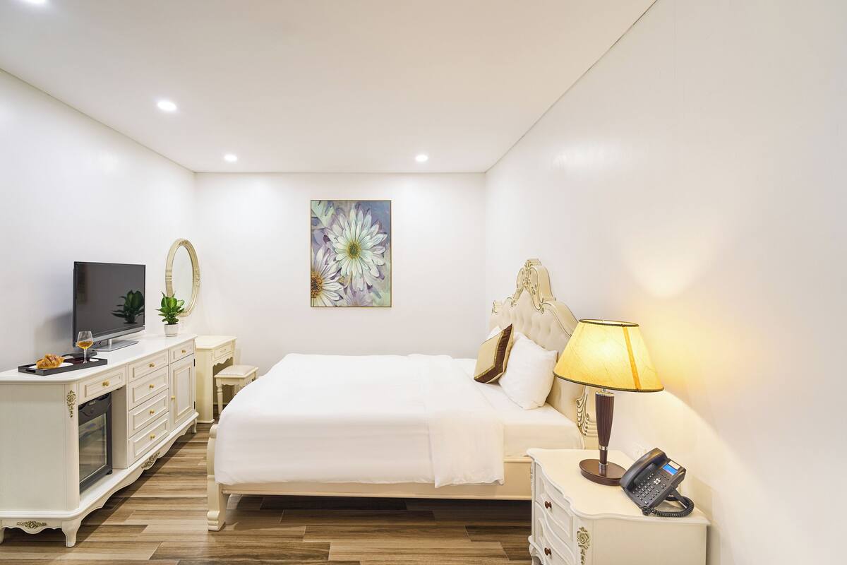 A hotel room with a bed, bedside table with a lamp, a telephone, a television, and a decorative mirror.