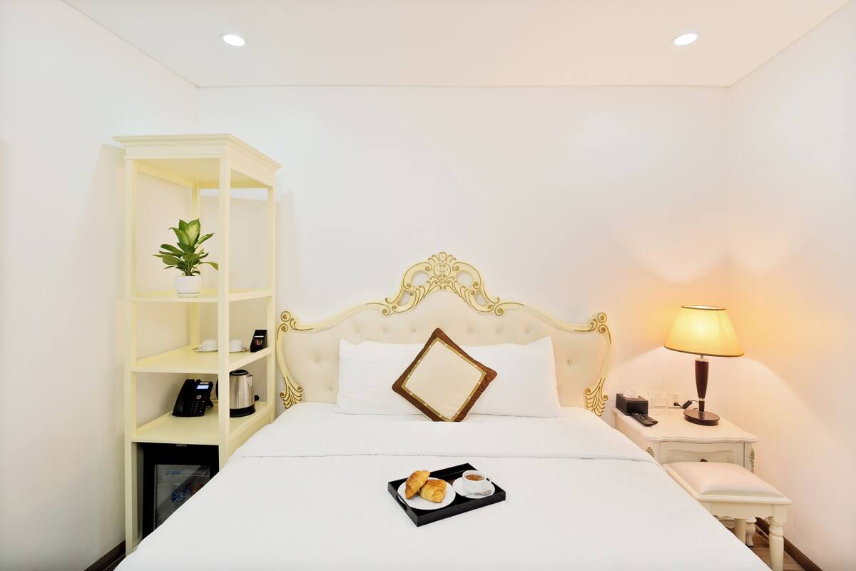 A neatly arranged hotel room with a bed, a bedside table with a lamp, a small shelf with a plant, and a tray with croissants and coffee.