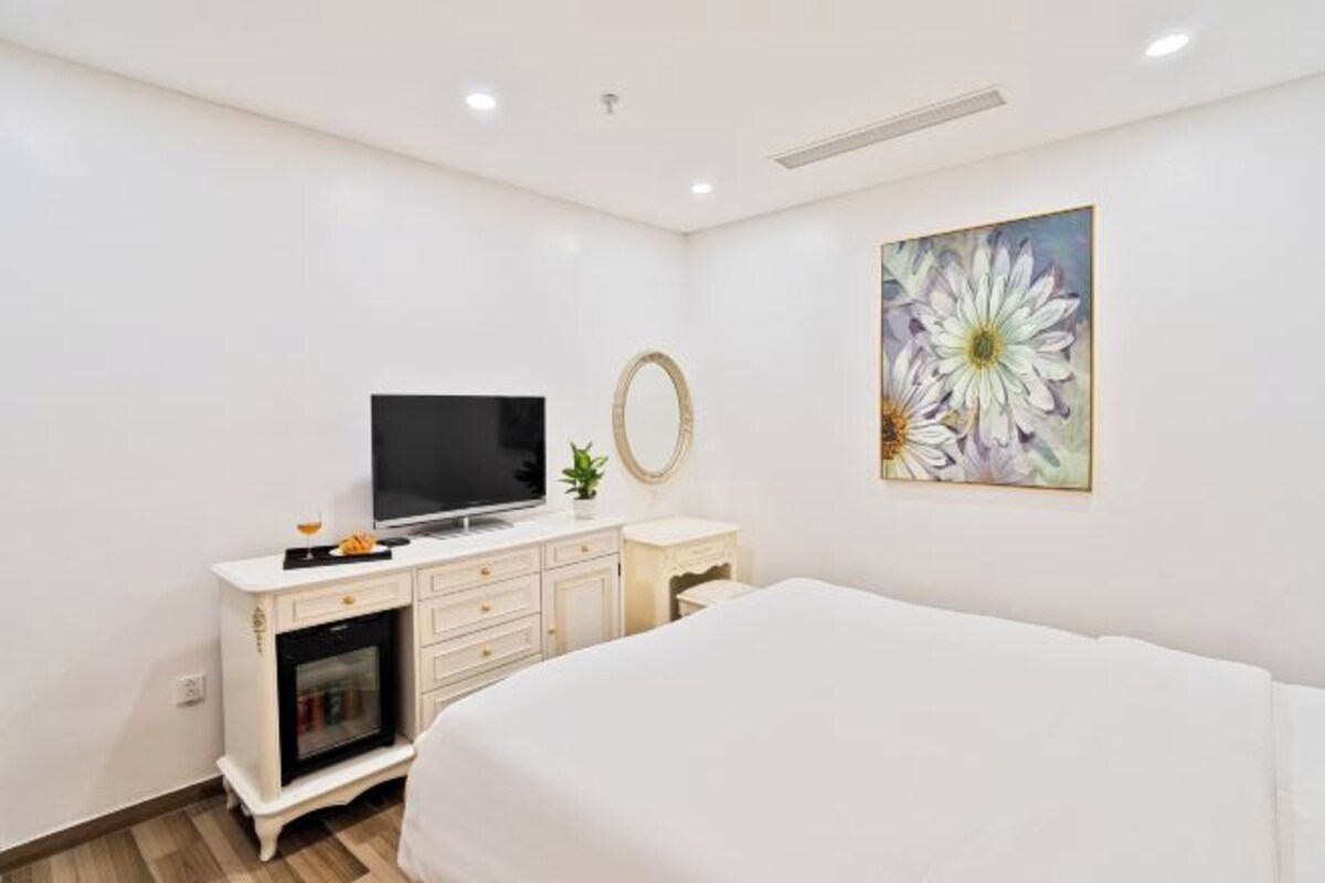Un dormitorio con una cama, televisión, cómoda blanca pequeña y chimenea, además de un cuadro con motivos florales en la pared.