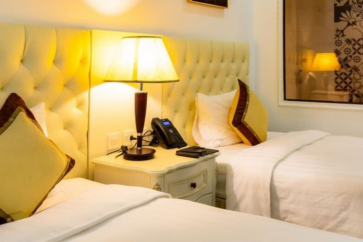 Una habitación de hotel con cabecera capitonada, dos camas con ropa blanca, una mesita de noche con lámpara, teléfono y un cajón pequeño.