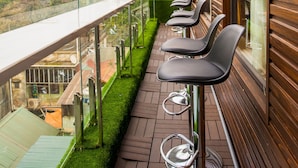 A balcony with a row of bar stools and a glass railing overlooking a green lawn and buildings.