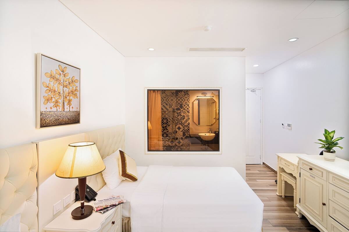 A modern hotel room with a large bed, a bedside table with a lamp, a mirror, and a view of the bathroom.