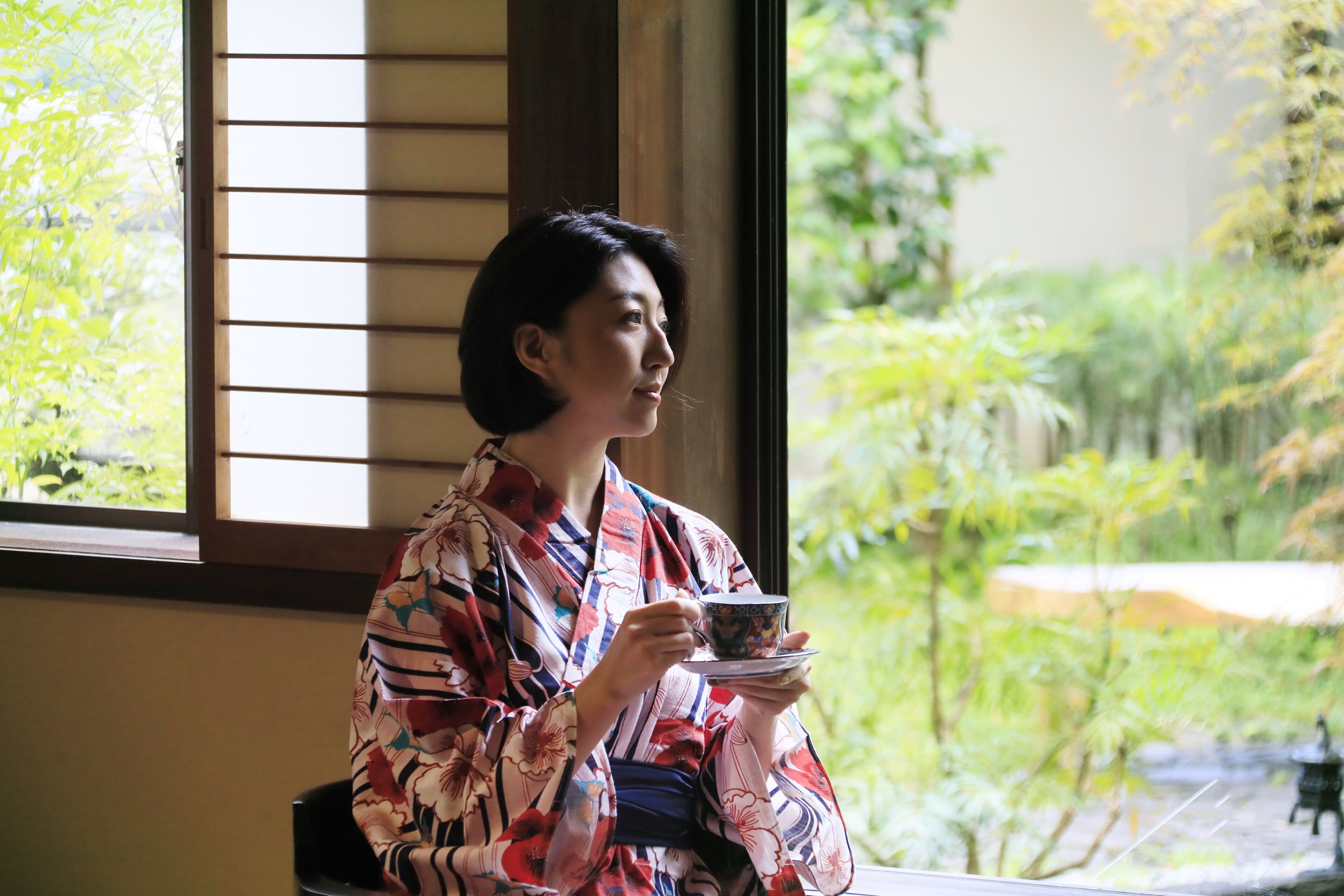 A person in a room with a view of greenery outside a window with blinds.