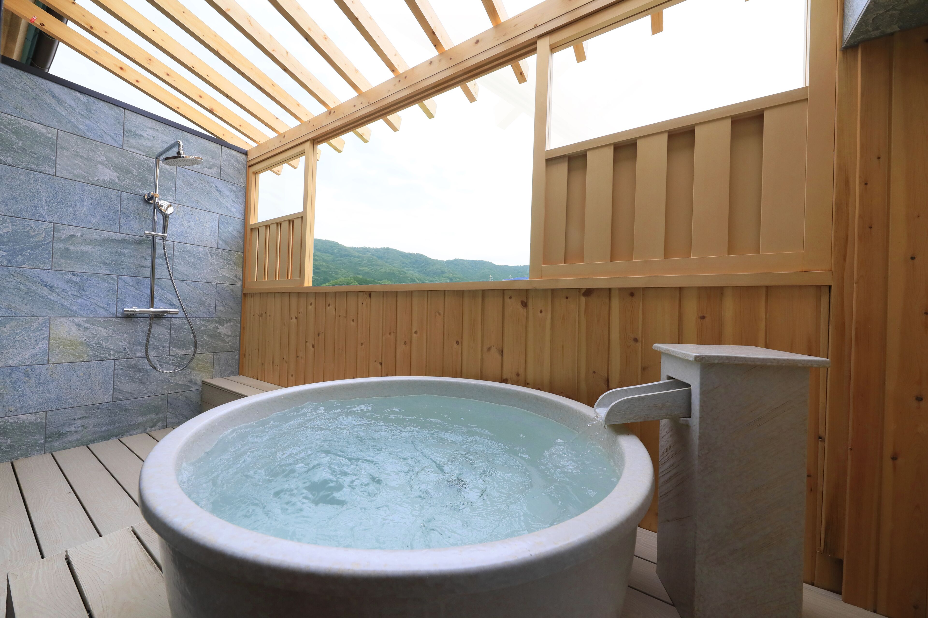 A hot tub with a wooden deck and stone walls, a showerhead, and a view of green hills.
