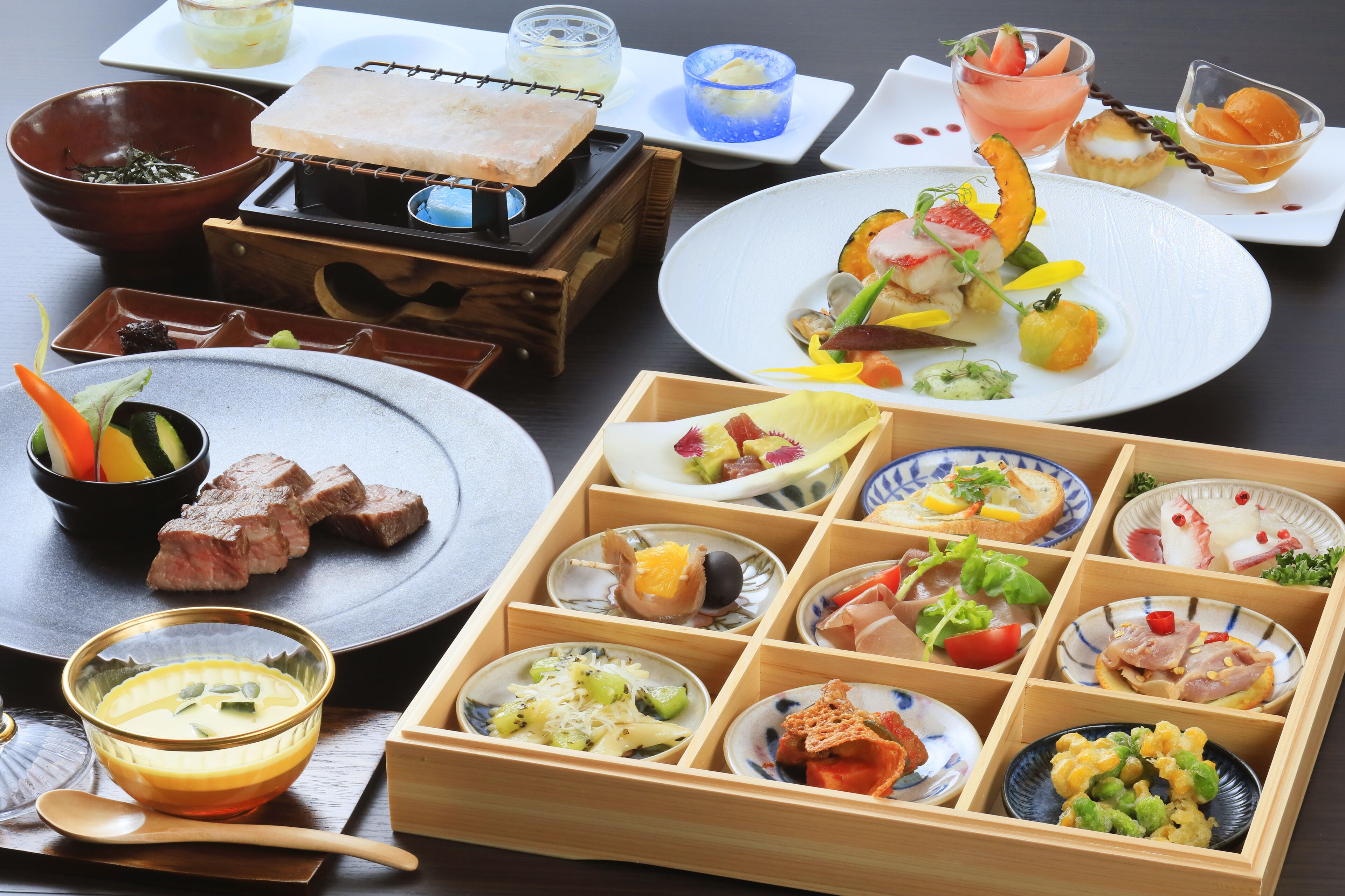 A variety of dishes and food items arranged on a table, including plates, bowls, and a wooden box with compartments.