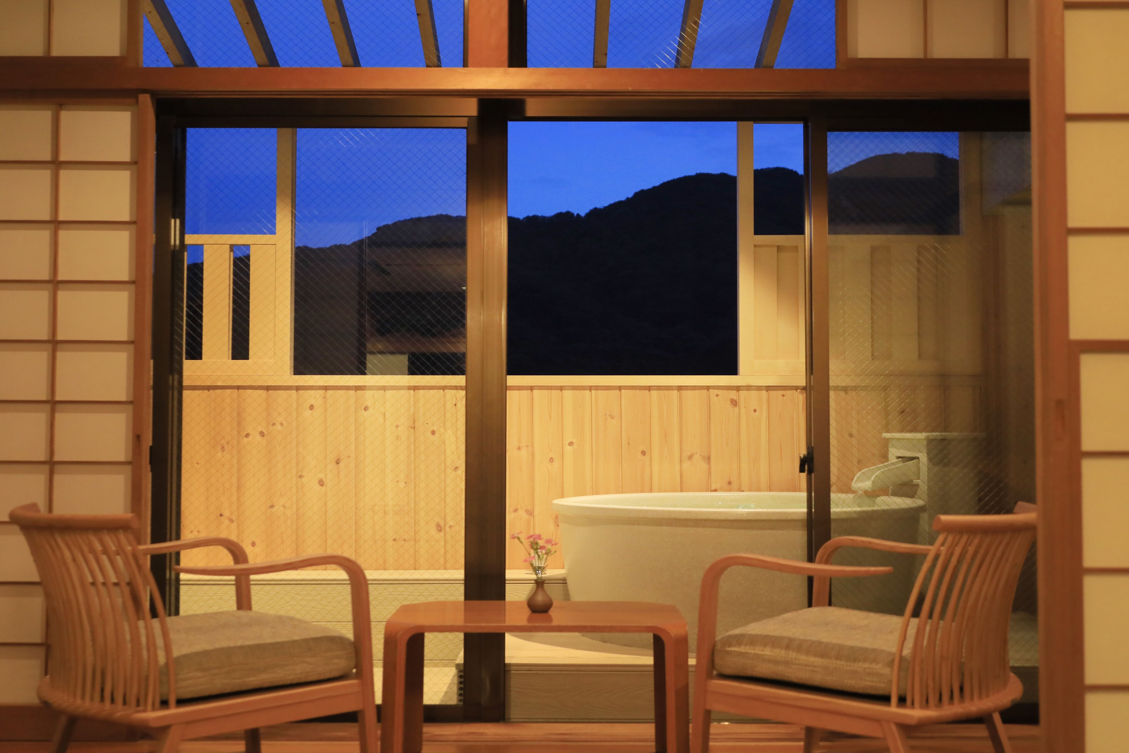 Una habitación con puerta corrediza de vidrio que da a una terraza de madera, un jacuzzi y dos sillas de mimbre.