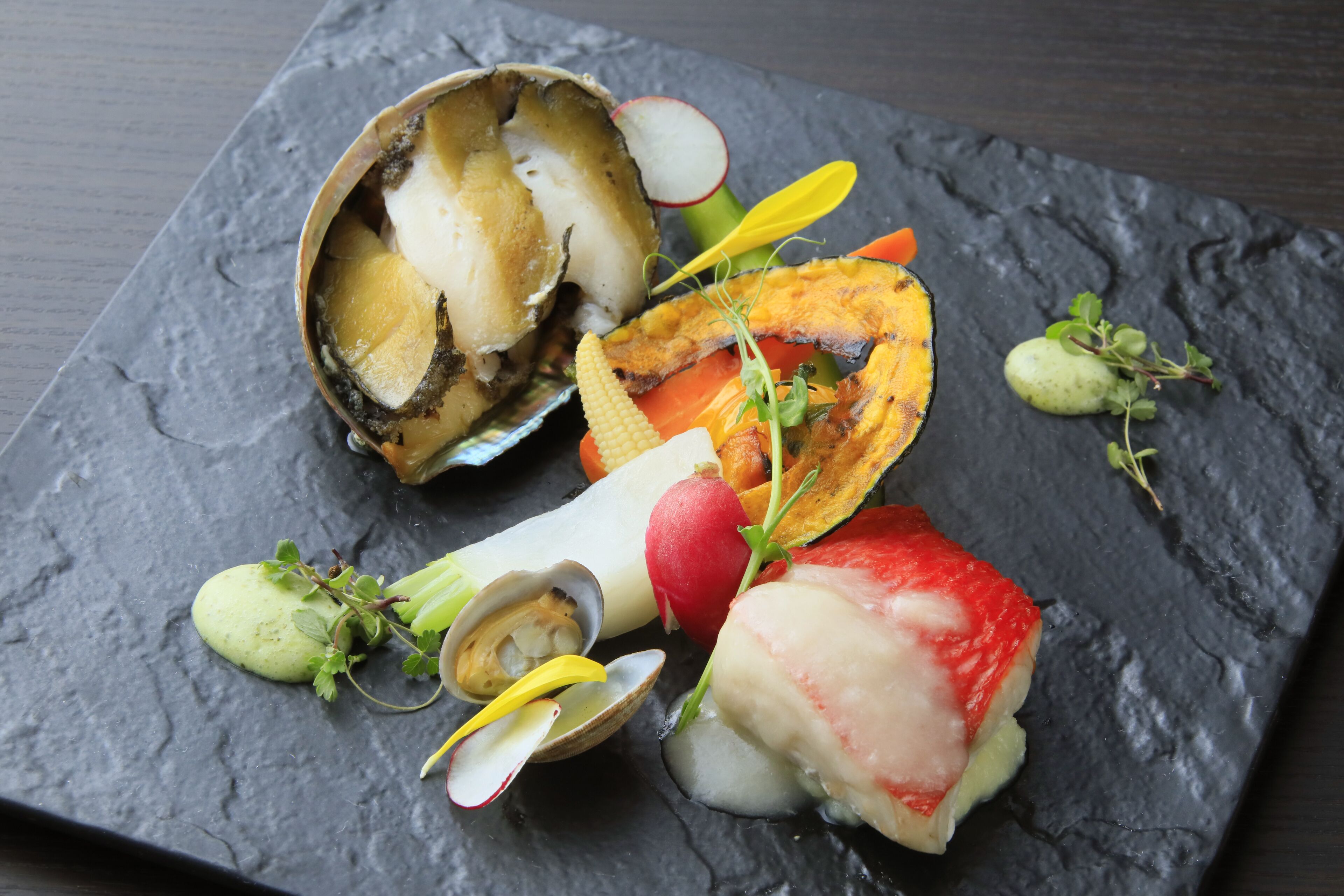 A square slate serving board with various seafood and vegetables, including clams, a radish, and a piece of fish.