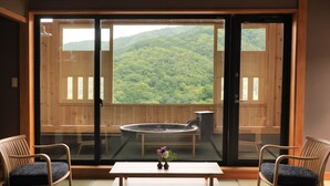 A room with a sliding glass door leading to a balcony with a hot tub and a view of a forested hillside. There are two wooden chairs and a small table with a vase of flowers. - Yugarawa Fuga (Yugawara)