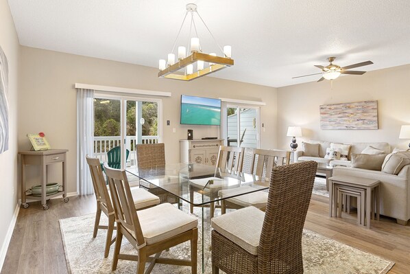 A spacious living area with a glass dining table, wicker chairs, a ceiling fan, and a television on the wall.