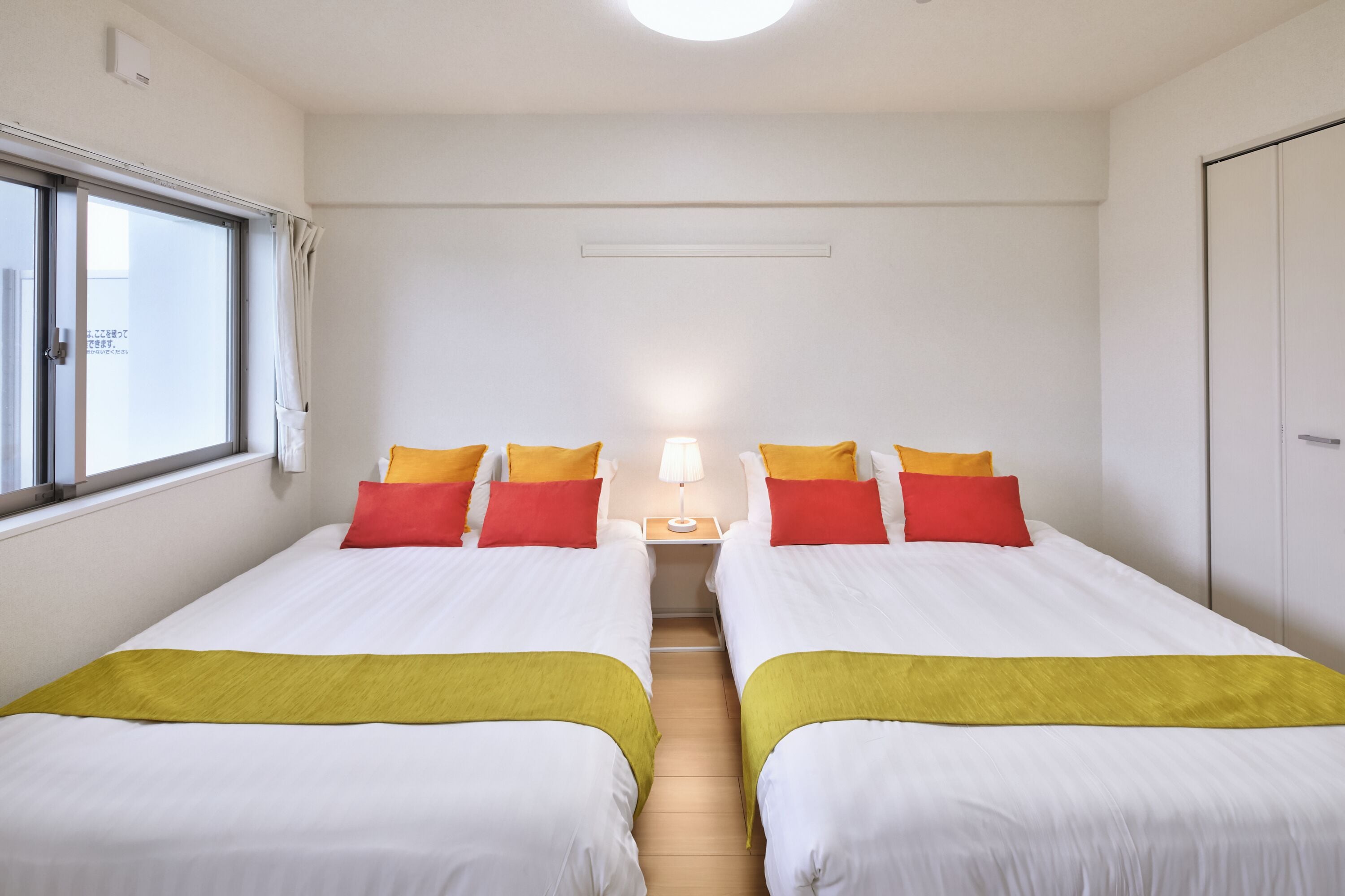 A hotel room with two beds, each with white bedding and yellow and red pillows. There is a nightstand with a lamp between the beds, and a window on the left side.