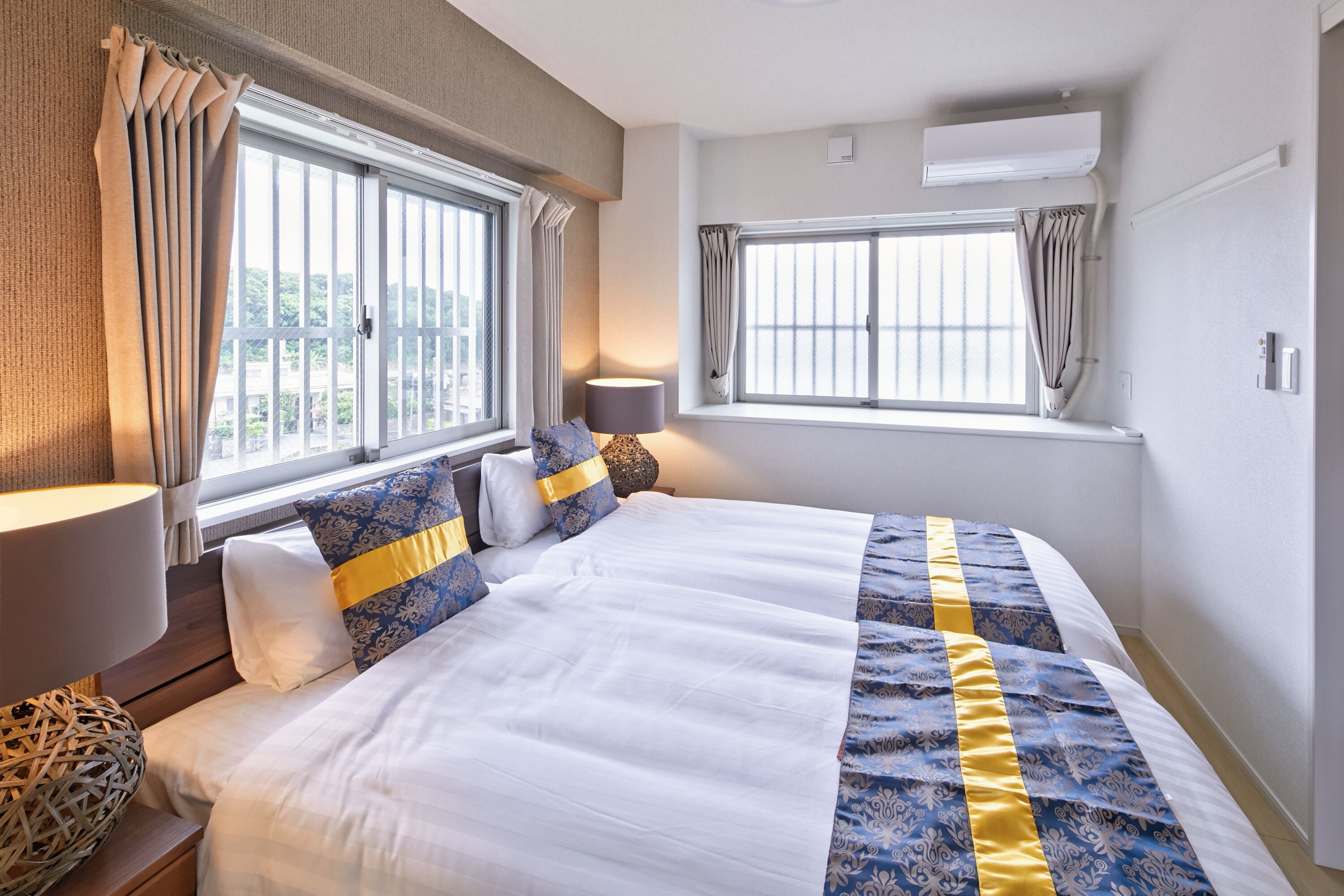 A hotel room with a large bed, two windows with curtains, a bedside lamp, and a wall-mounted air conditioner.