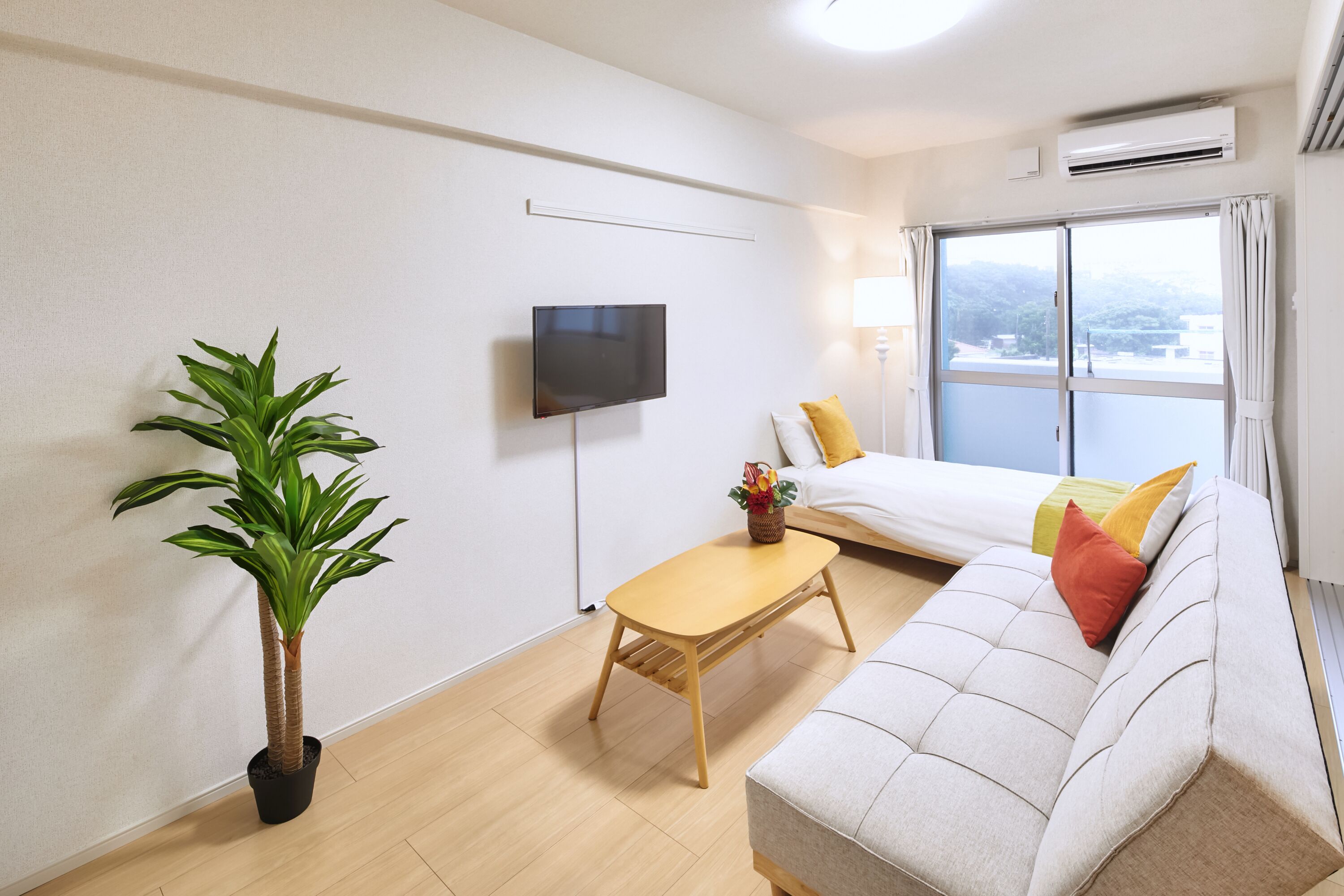 A modern hotel room with a sofa, a flat-screen TV, a small table, a potted plant, and a large window with curtains.