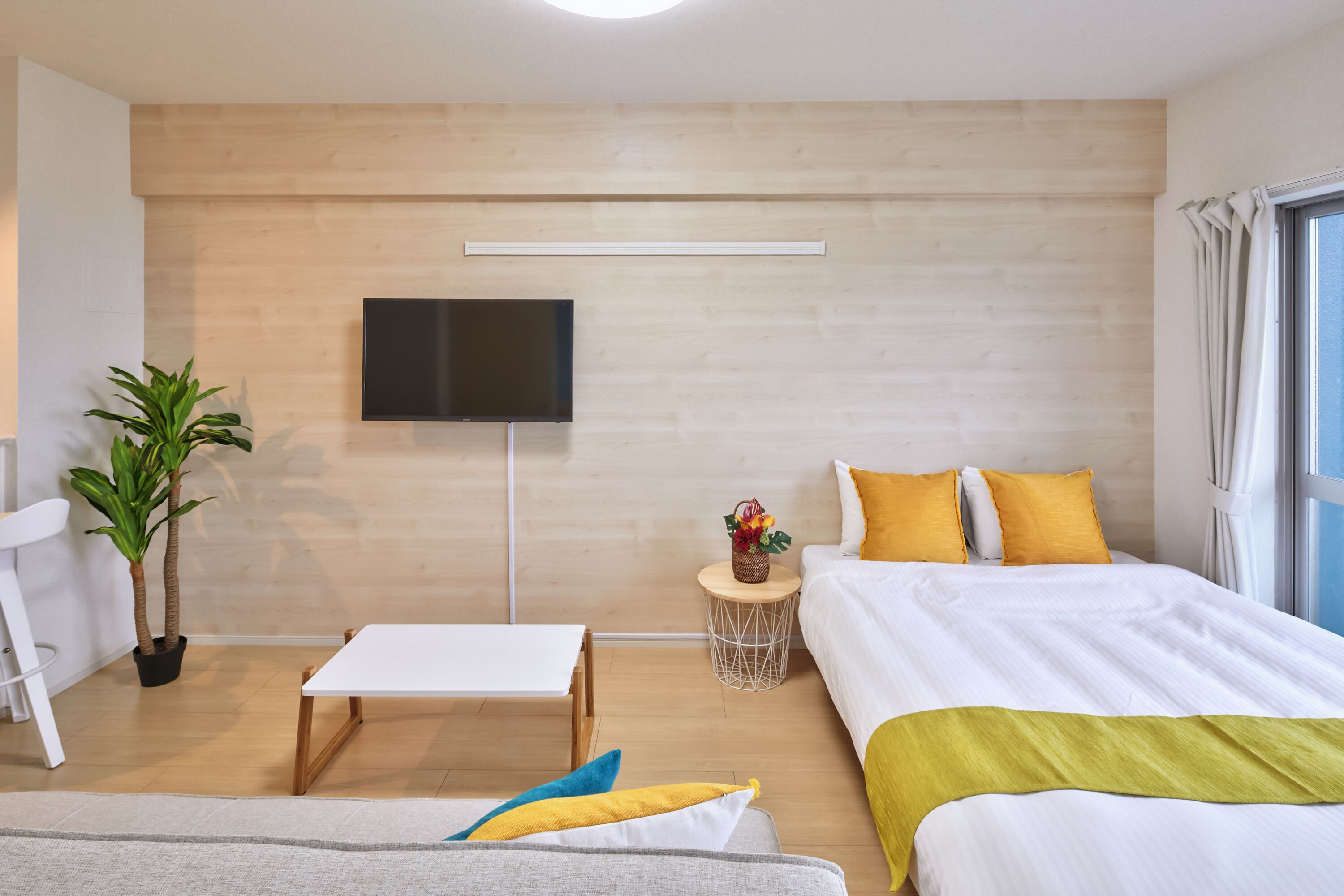 Una habitación de hotel moderna con una pared de madera, un televisor de pantalla plana, una cama con ropa de cama amarilla y blanca, una pequeña mesa redonda con una planta y una silla blanca.