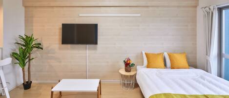 Una habitación de hotel moderna con una pared de madera, un televisor de pantalla plana, una cama con ropa de cama amarilla y blanca, una pequeña mesa redonda con una planta y una silla blanca.