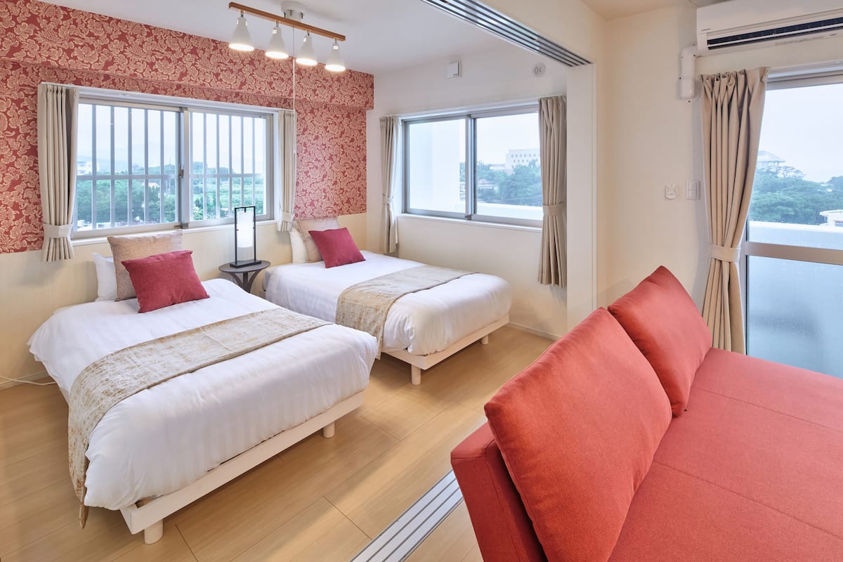A hotel room with two beds, a red sofa, and large windows with curtains.