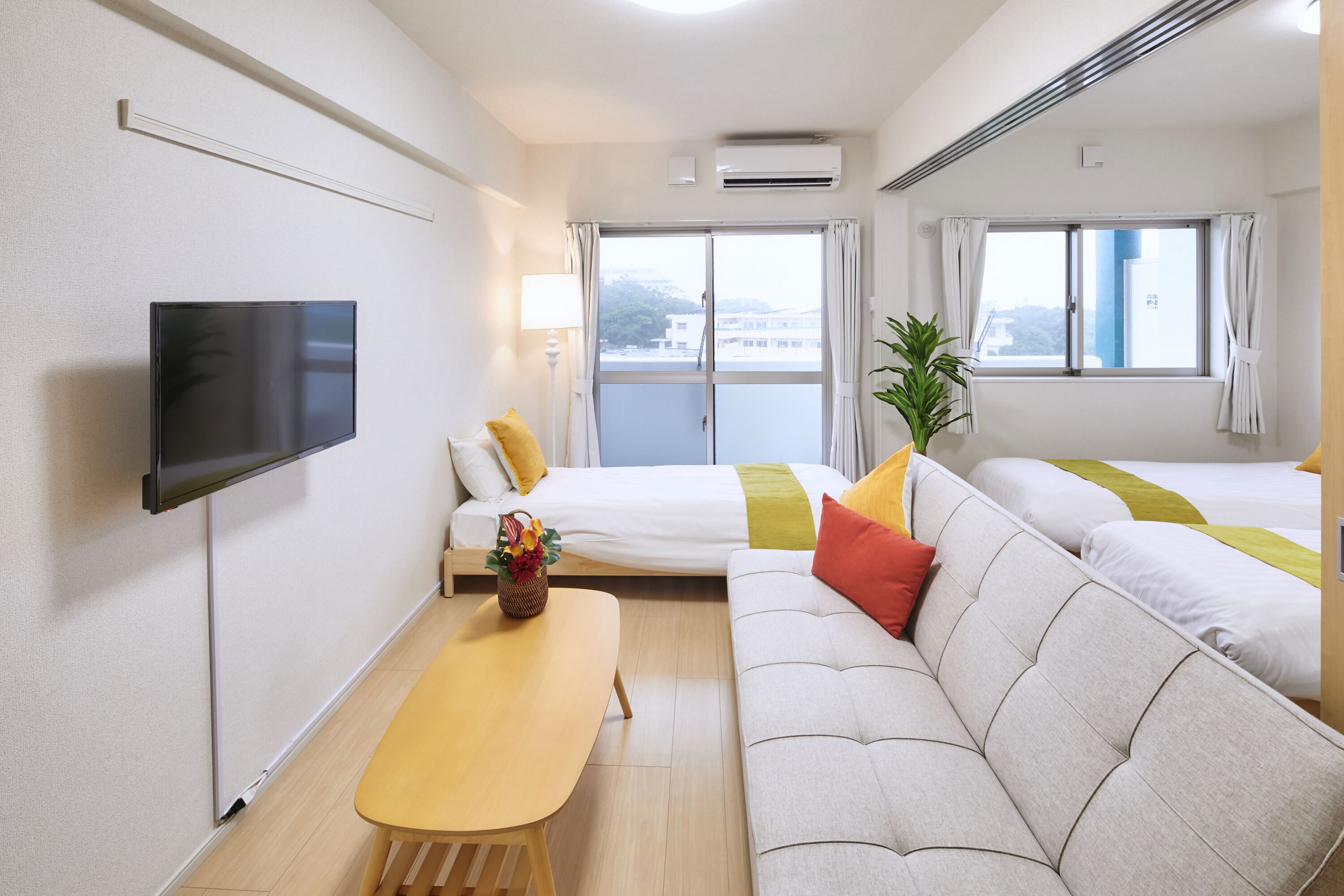 A hotel room with a flat-screen TV, a sofa, a bed, a small table with a vase, and a window with curtains.