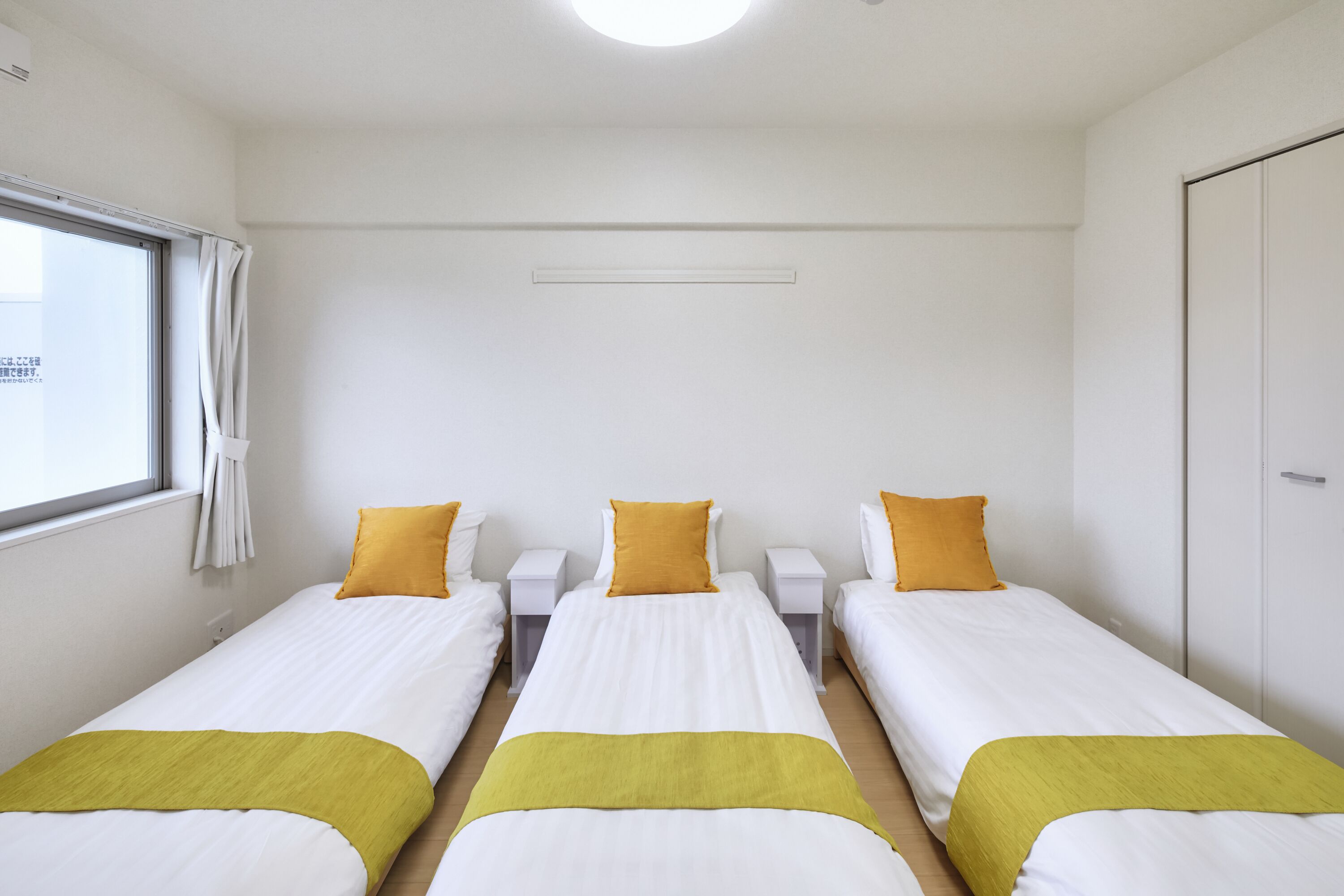 A hotel room with three single beds, each with a white and yellow bedspread, and a window with curtains.