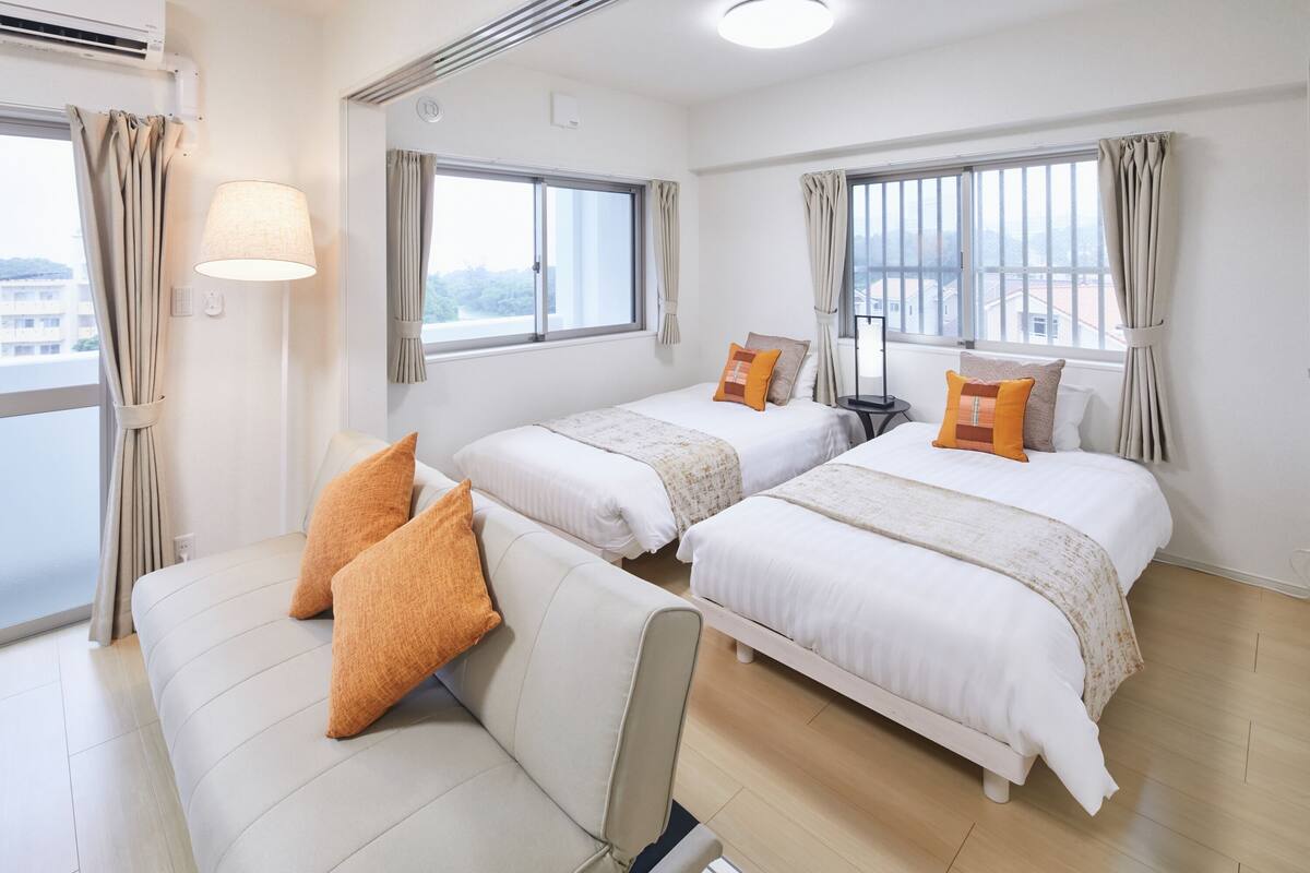 A hotel room with two single beds, a sofa, a lamp, and a view of the city.