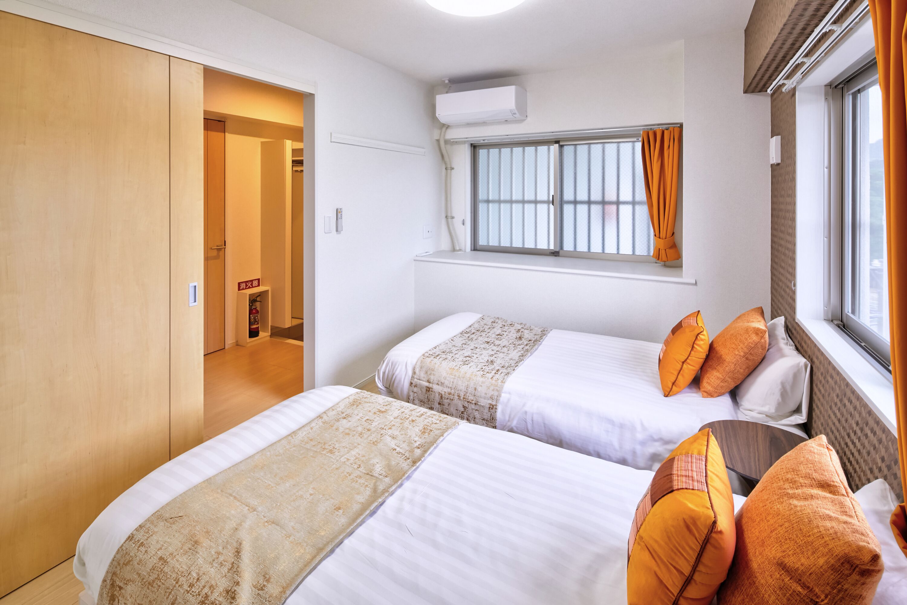 Ein Hotelzimmer mit zwei Betten, einem Fenster mit orangefarbenen Vorhängen, einer Decken-Klimaanlage und einem Holzschrank.