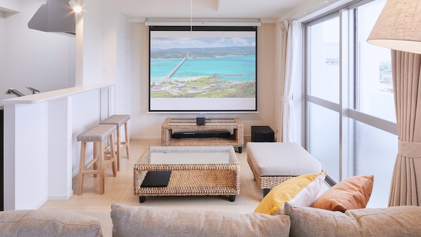 Una sala de estar moderna con una amplia pantalla de televisión mostrando una vista panorámica, mobiliario de mimbre y una cocina con taburetes.