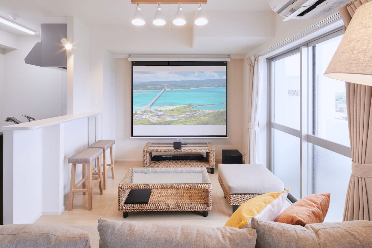 A modern living room with a large TV screen displaying a scenic view, wicker furniture, and a kitchen area with stools.