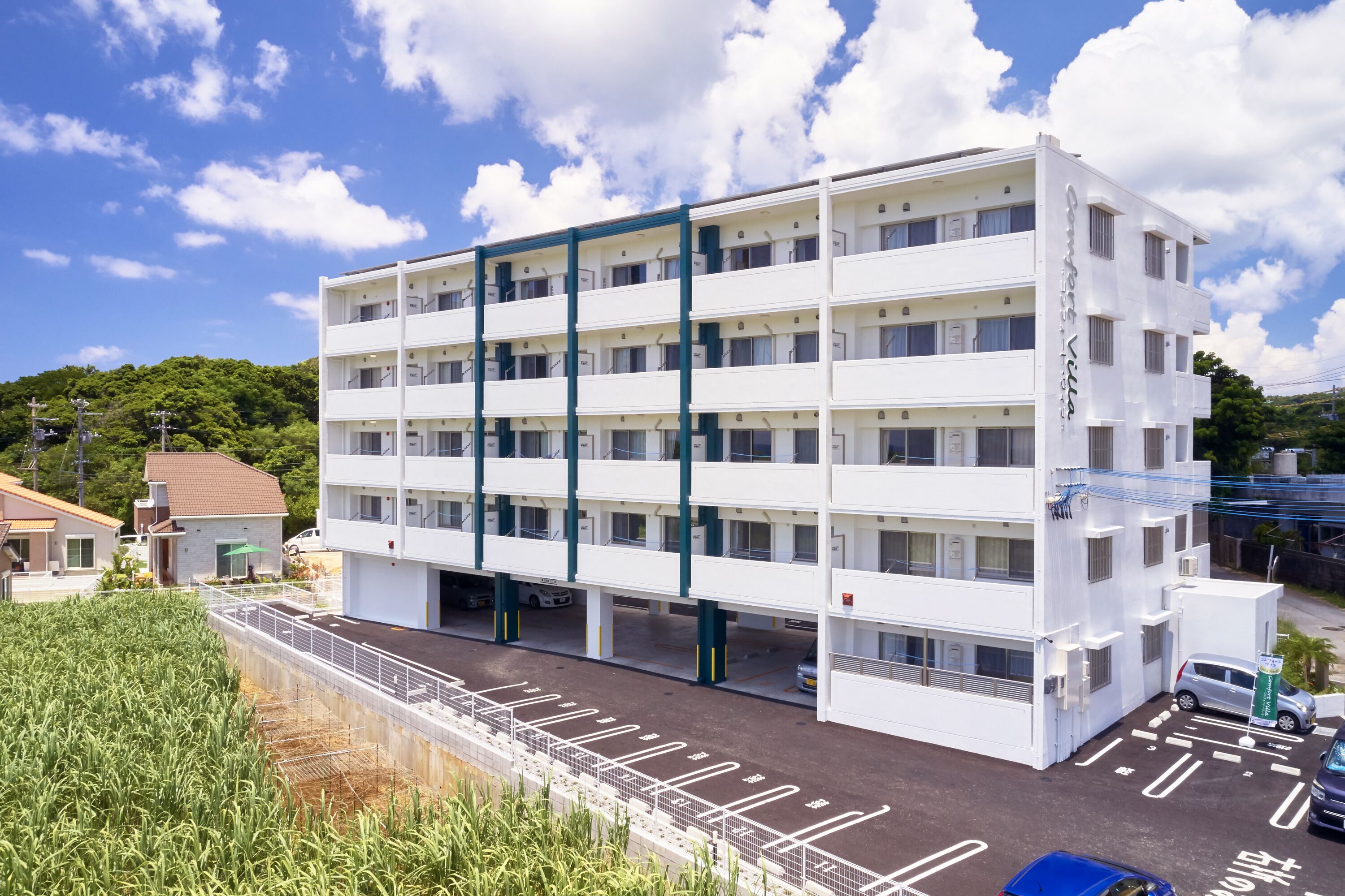 A multi-story hotel with a white facade and green accents, featuring a parking area and a view of surrounding greenery.