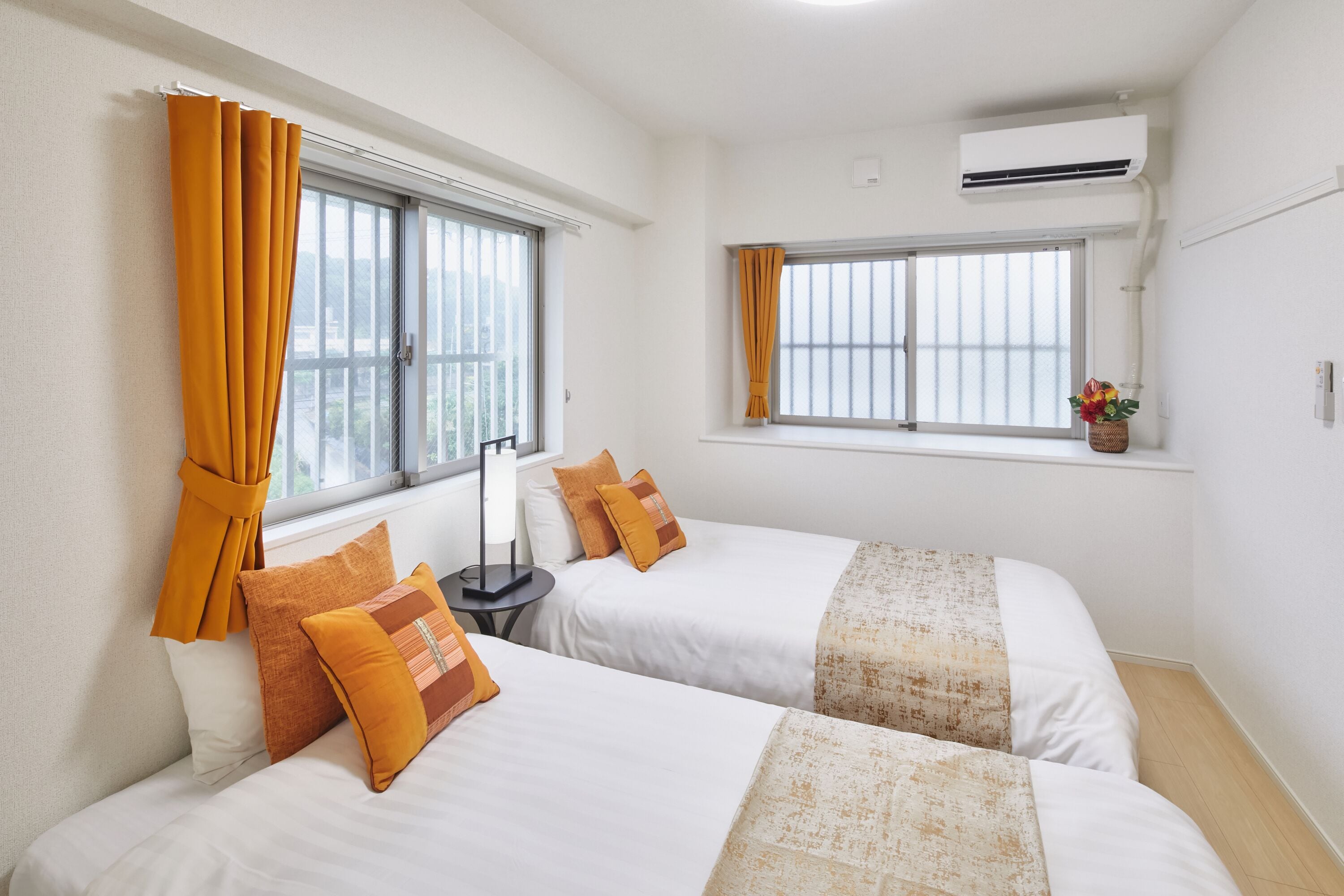 A hotel room with two beds, a window with curtains, an air conditioning unit, and a small table with a lamp.