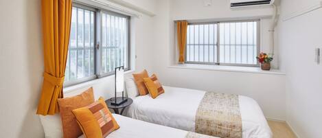 A hotel room with two beds, a window with curtains, an air conditioning unit, and a small table with a lamp.