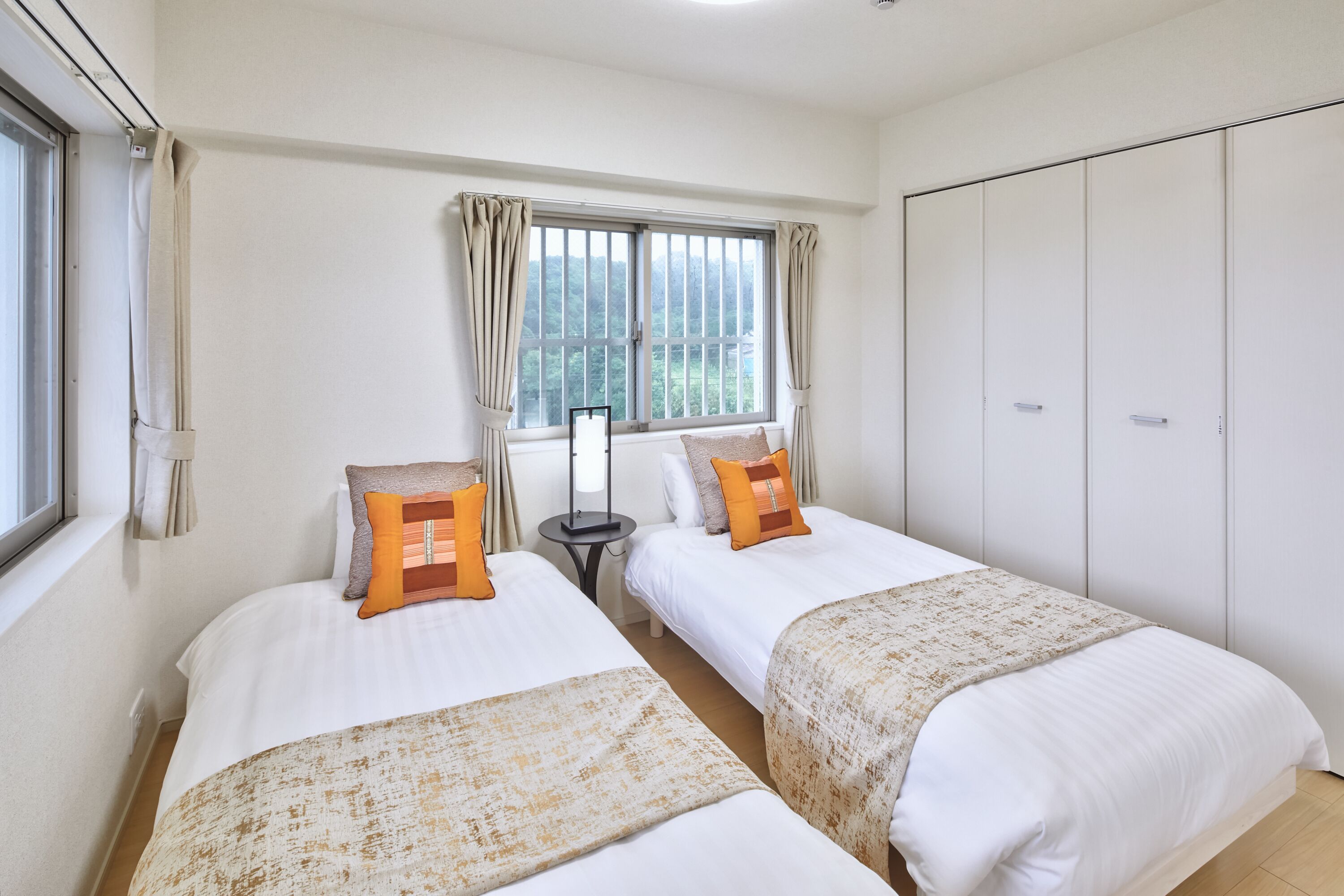 Habitación de hotel con dos camas, una ventana con cortinas, una mesita de noche con lámpara y un armario.