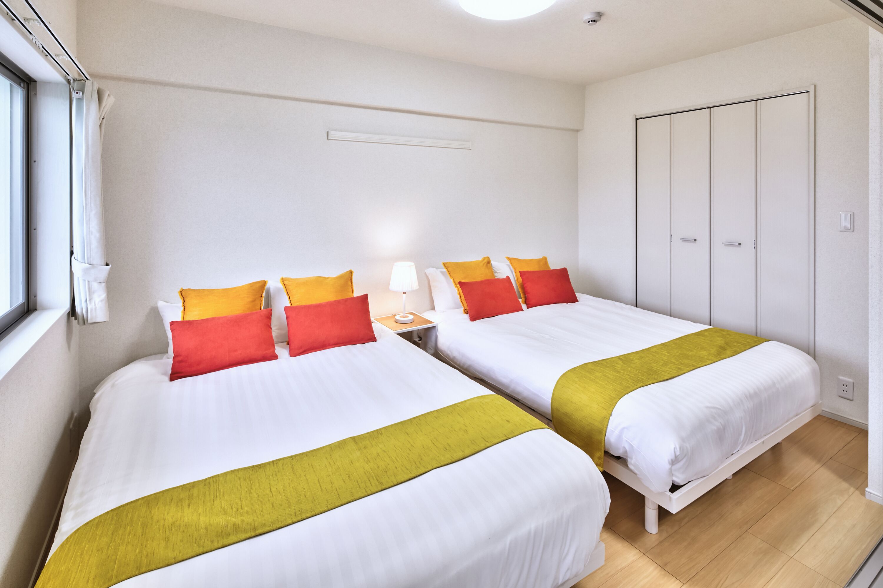 Habitación de hotel con dos camas, cada una con ropa blanca y almohadas de colores, una mesita de noche con lámpara y un armario de dos puertas.