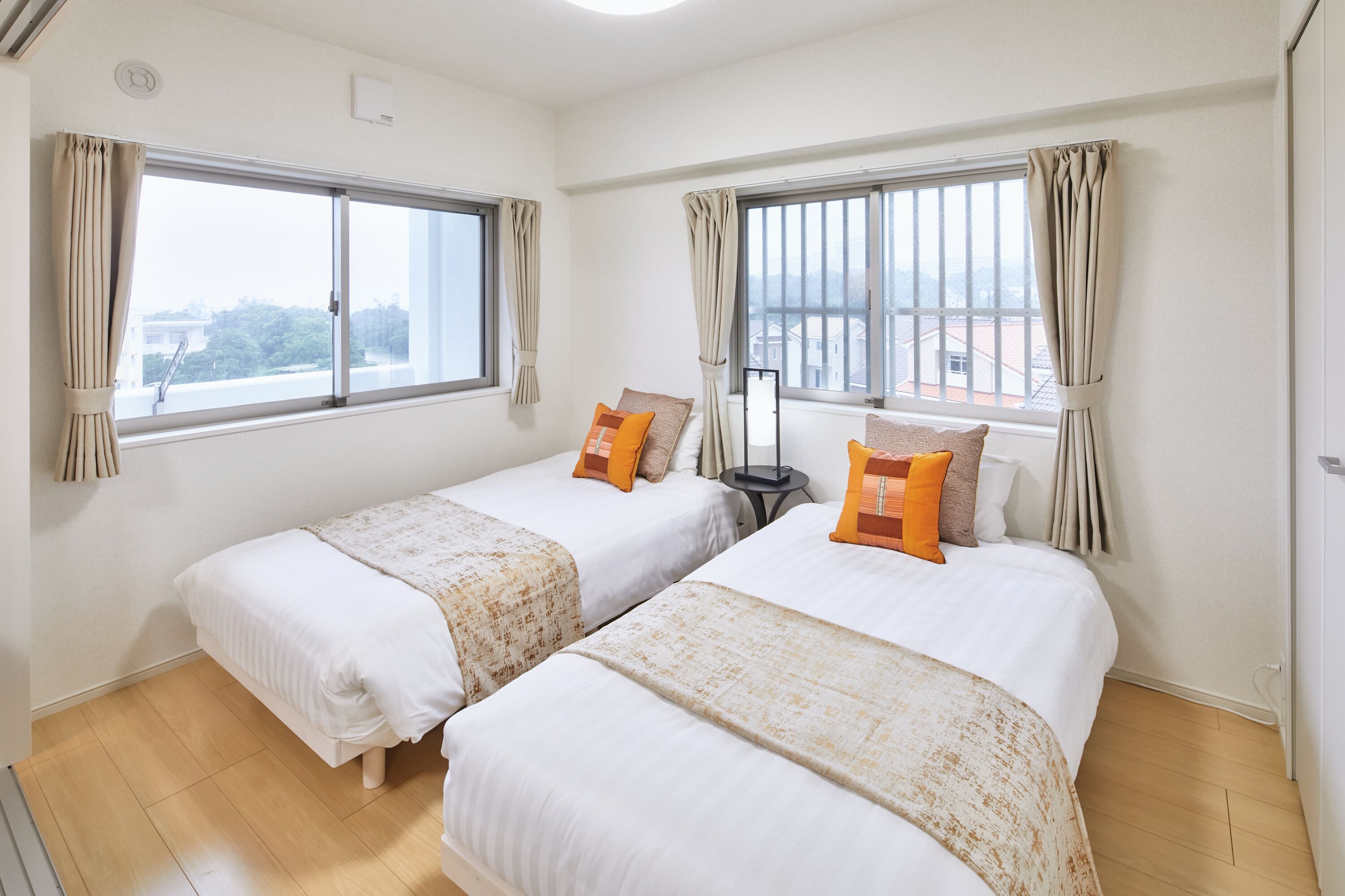 Ein Hotelzimmer mit zwei Einzelbetten, beigen Vorhängen, einem kleinen Tisch und Blick auf Gebäude durch die Fenster.