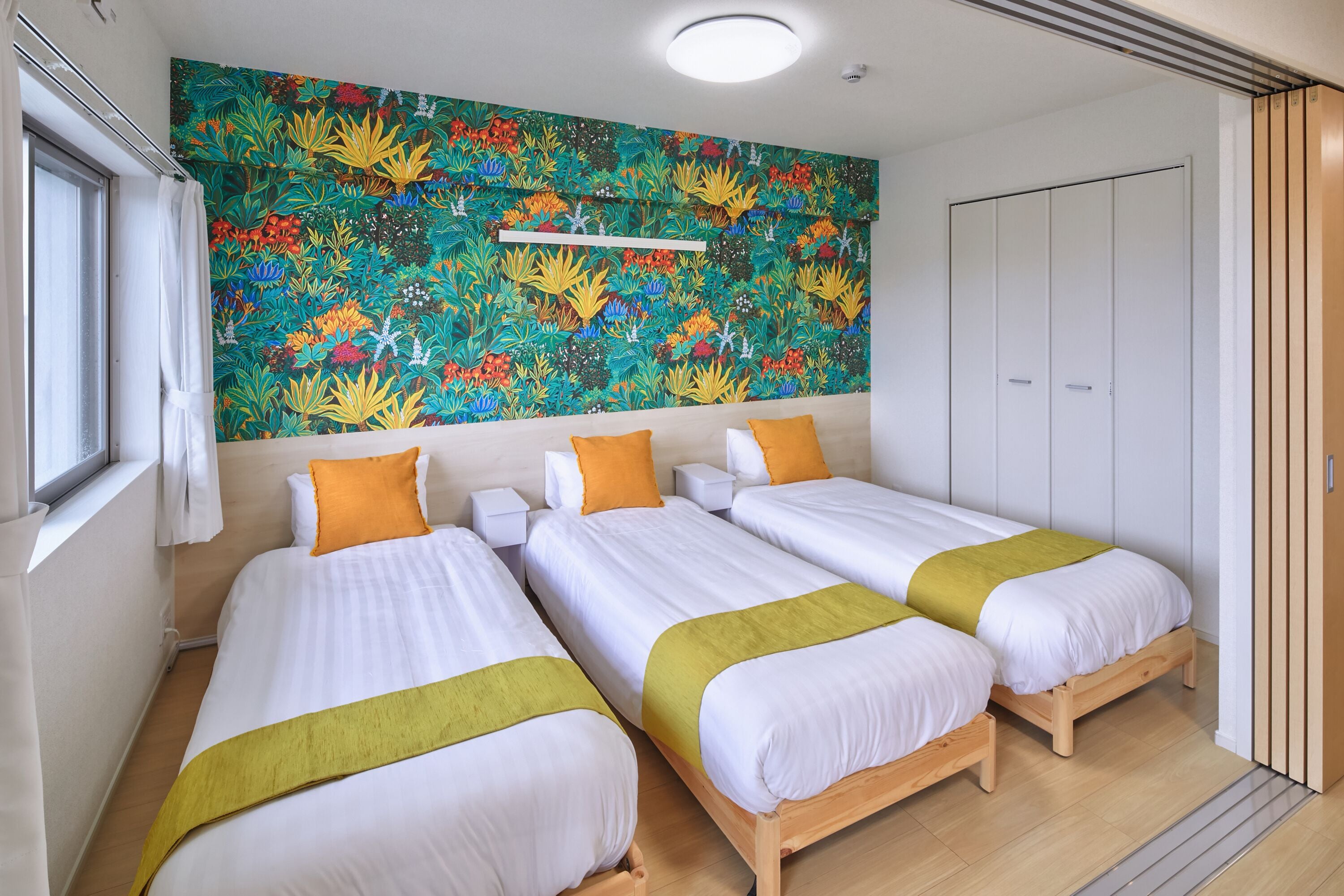 Habitación de hotel con dos camas, un mural floral de colores, un armario blanco y una ventana con cortinas blancas.