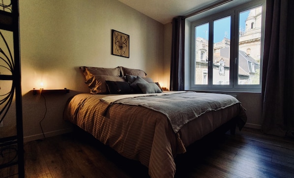 A bedroom with a large bed, a window with a view of buildings, and a wall-mounted light fixture.