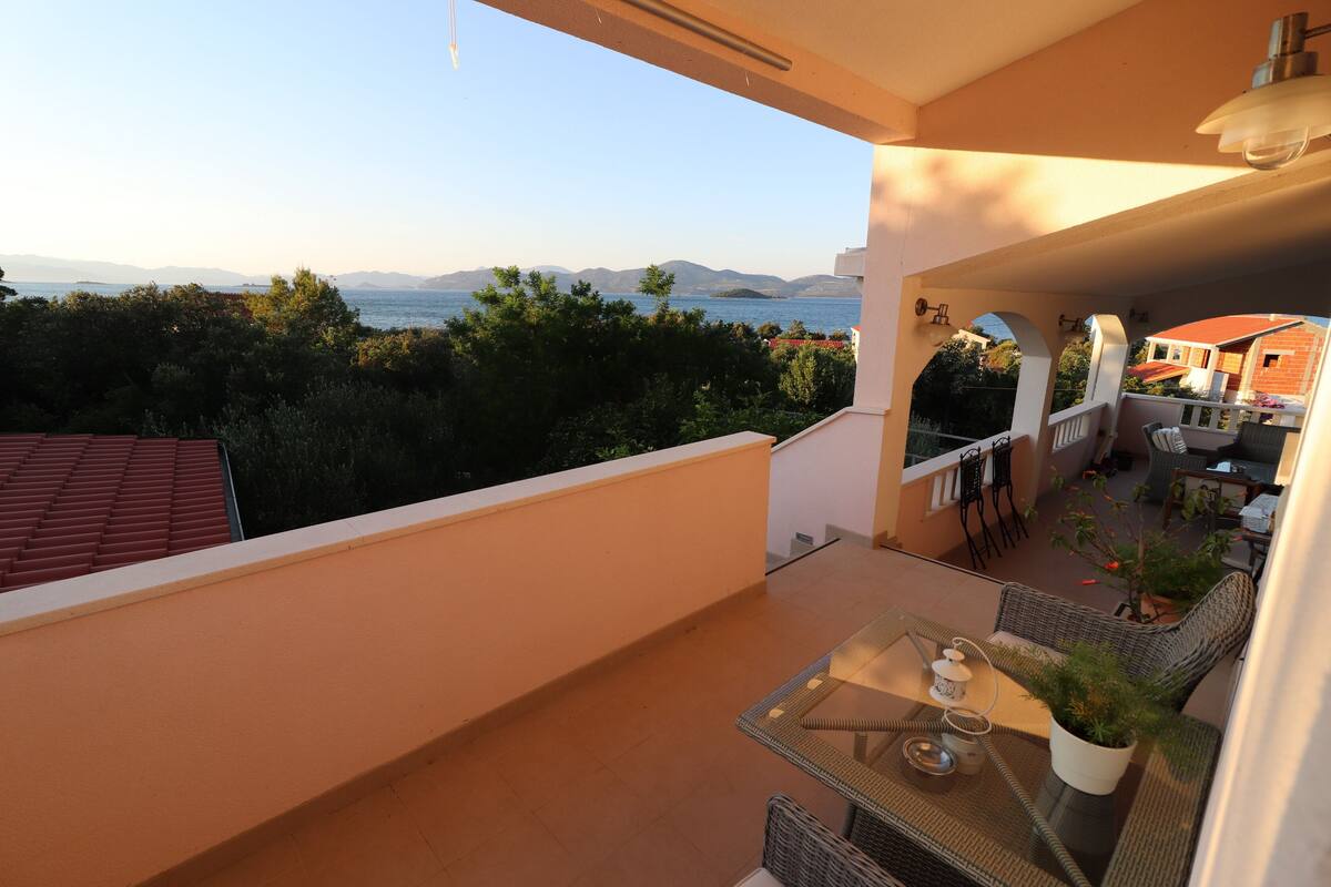 A balcony with a table and chairs, a potted plant, and a view of the sea and mountains.