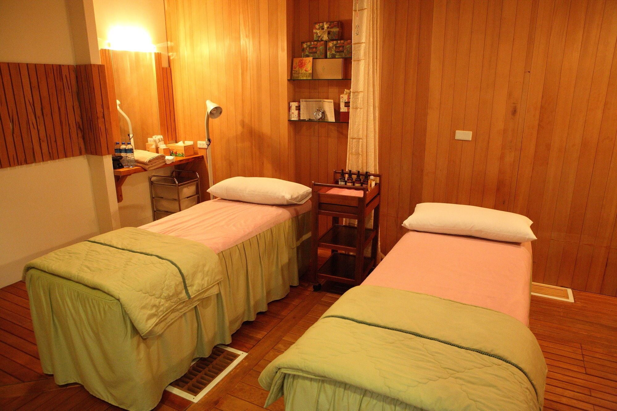 A room with two single beds, wooden walls, a small desk with a lamp, and a shelf with toiletries.