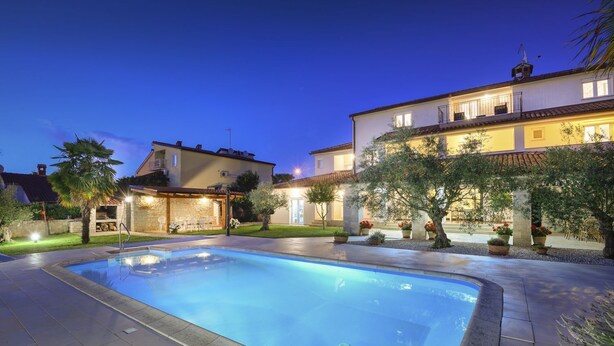 A swimming pool with a stone deck, surrounded by a well-maintained garden and a large house with multiple balconies.