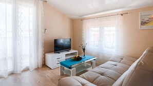 A living room with a sofa, a TV on a white cabinet, a glass coffee table with a vase and fruit, and sheer curtains.