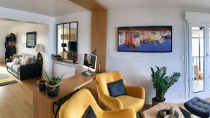 En rummelig opholdsstue med sofa, lænestol og spisebord. Et stort vindue giver naturligt lys og udsigt til det fri.