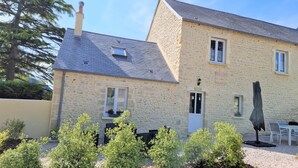 Un bâtiment en pierre doté d’un toit en pente, d’une porte avec une fenêtre et d’un petit jardin agrémenté d’arbustes.