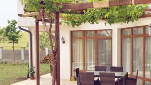A covered patio with wicker furniture, a pergola with vines, and a view of a house with large windows. - Generaator House (Pomorie)
