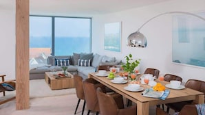 A modern living room with a large window, a dining area with a wooden table set for breakfast, and a cozy seating area with a sofa and cushions. - Beachfront Townhouse, Lovely Sea Views, Newquay (Newquay)
