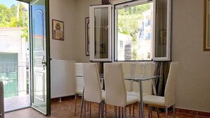 Zona pranzo con tavolo in vetro e sedie bianche, un termosifone e vista su una zona residenziale attraverso ampie finestre.