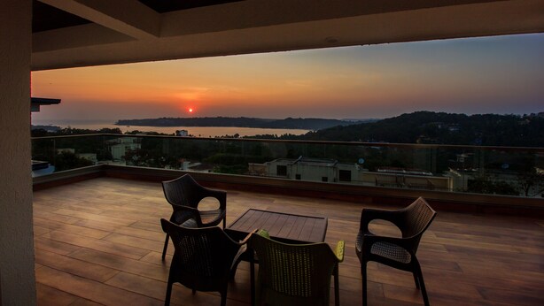 A balcony with two chairs and a table, overlooking a sunset over a cityscape. - ROSASTAYS North Goa Rosa Kuber Mandovi (Alto de Porvorim)