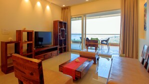 A spacious living room with a flat-screen TV, wooden furniture, a balcony with a view, and a ceiling fan. - ROSASTAYS North Goa Rosa Kuber Mandovi (Alto de Porvorim)