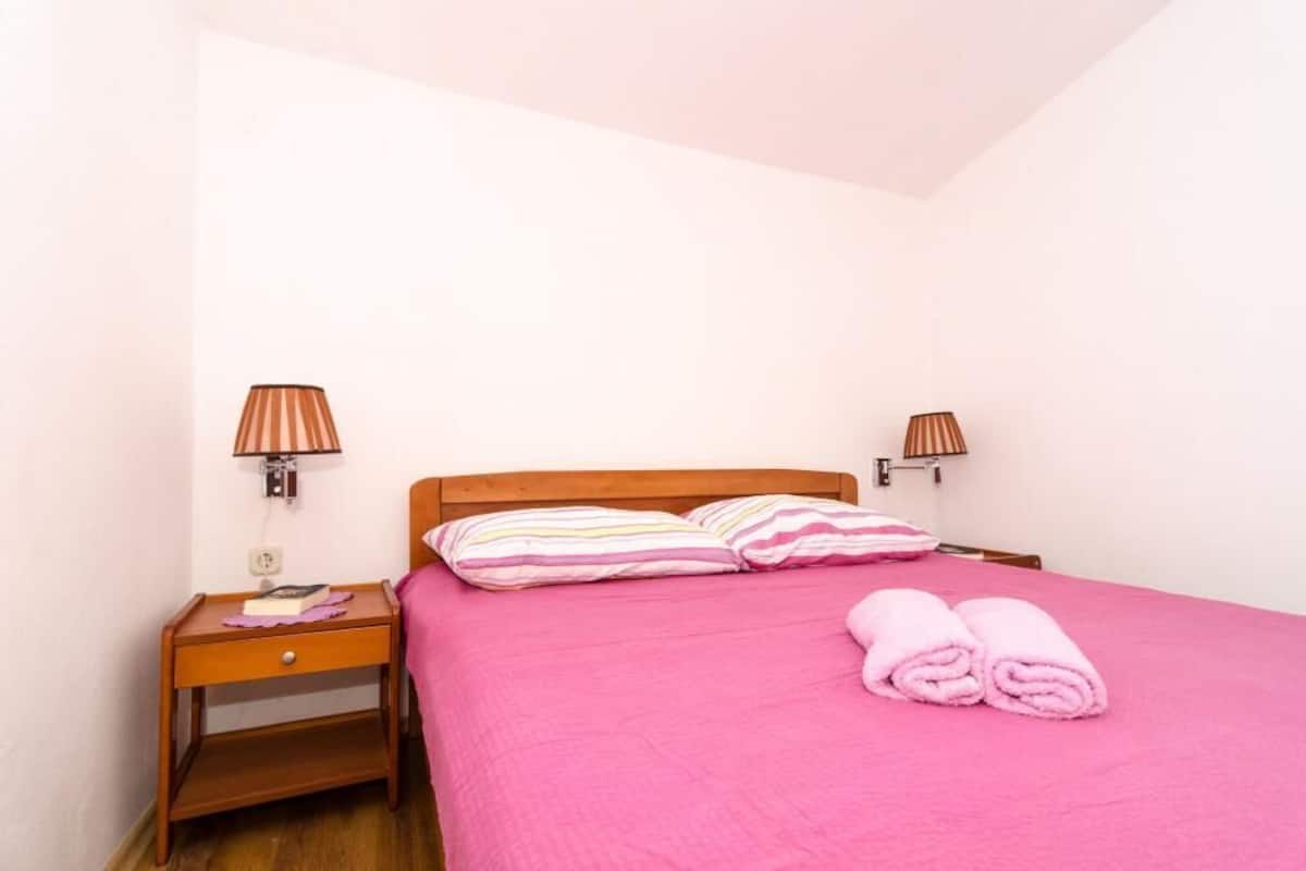 A bedroom with a wooden headboard, two bedside lamps, a pink bedspread, striped pillows, a nightstand with a book and a lamp, and rolled towels on the bed.