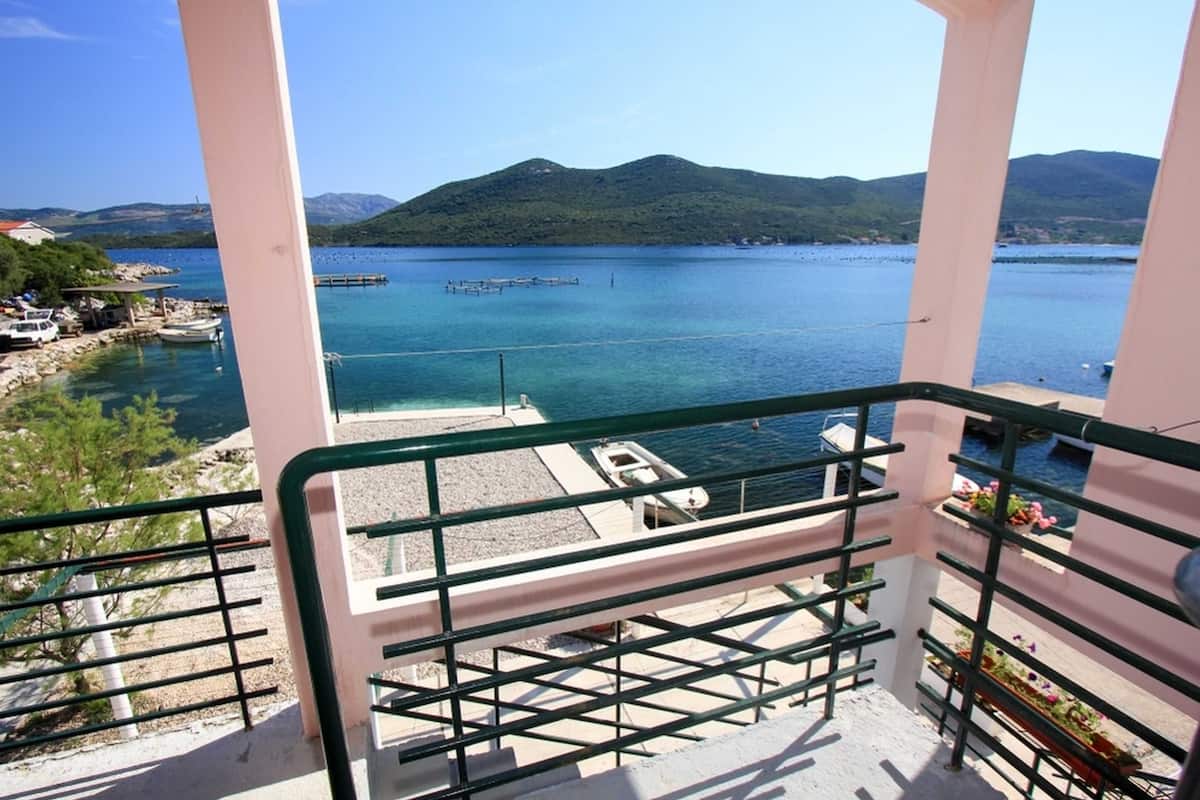 A balcony with a view of a calm sea, a dock, and distant mountains.