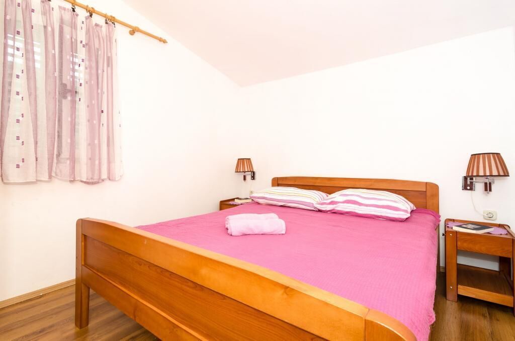 Un llit individual amb estructura de fusta i un llençol de llit rosa, dues taules de nit amb làmpades, i una finestra amb cortines.